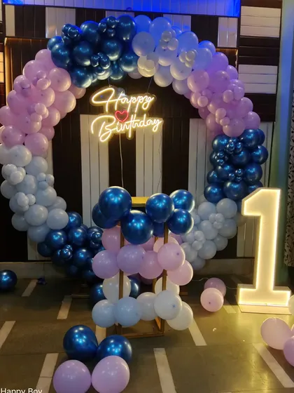 A circular balloon arch for a first birthday in shades of blue, purple, and pink. The setup is completed with a "Happy Birthday" neon sign and a large light-up number "1".