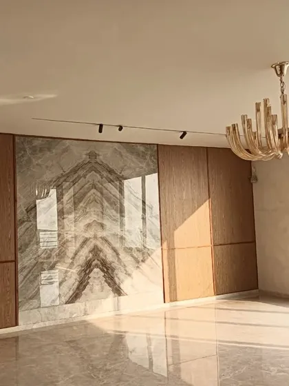 This penthouse living room is nearing completion, and the key elements are already shining. The feature wall combines bookmatched marble with warm wood paneling, creating a luxurious and symmetrical focal point. Natural light from the large window illuminates the polished marble floors, enhancing the sense of space and elegance.