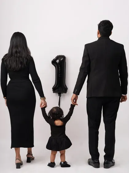 A creative family photo taken from behind, showing parents and their daughter holding hands with a '1' balloon to celebrate her first birthday.