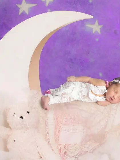 A sweet newborn portrait on a moon prop against a starry purple night sky backdrop.