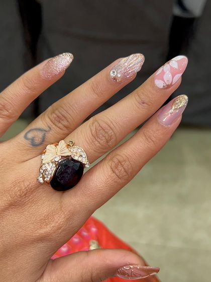 A soft and glamorous set with a mix of textures and finishes. It includes pink floral nails, glitter ombré, gold foil, and sculpted shell details for a multi-dimensional and feminine look.