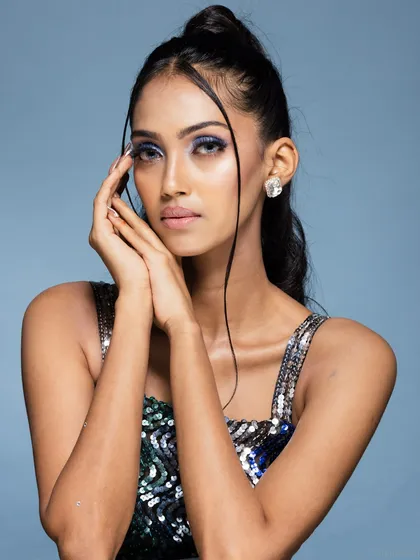 A soft beauty portrait featuring a model with her hands framing her face. The focus is on the shimmering blue makeup and delicate jewellery.