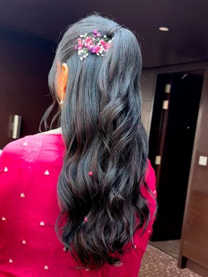 A soft and pretty half-up ponytail for a Haldi or Mehendi ceremony. The style is adorned with a small cluster of fresh baby's breath and pink flowers, adding a delicate, romantic touch.