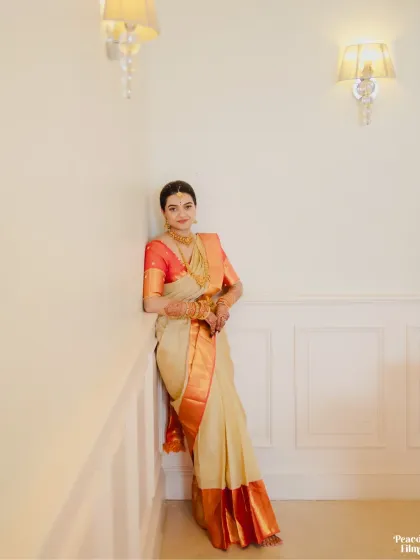 A full-length portrait of the bride in her classic South Indian wedding saree, leaning against a wall with a gentle smile.