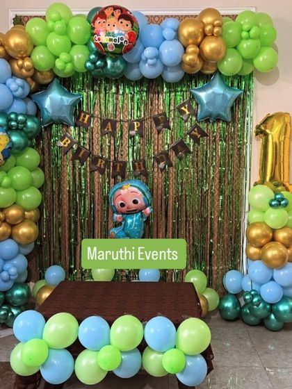 A Cocomelon-themed first birthday party. The decor features a balloon arch in the show's signature green, blue, and gold, with a character cutout and a shiny green foil backdrop.