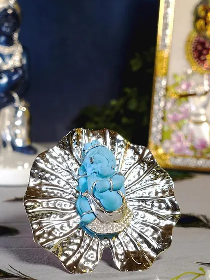 A serene Bal Krishna, carved from a beautiful blue stone, rests on a silver-plated lotus leaf. This idol combines natural elements with fine metalwork, symbolizing purity and divine love.