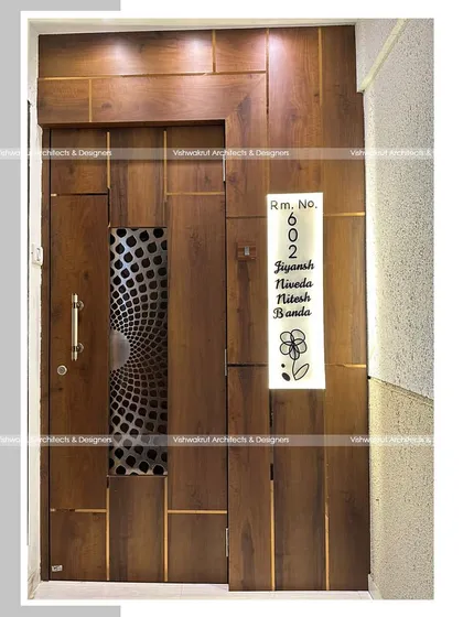 A modern entrance door design for a 1BHK apartment. The door features a unique metal jali insert and is surrounded by wooden paneling with brass inlays, creating a sophisticated and contemporary look.