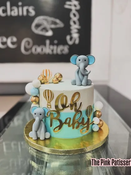 An "Oh Baby" shower cake with cute elephant toppers and hot air balloon details in a blue, white, and gold color scheme.