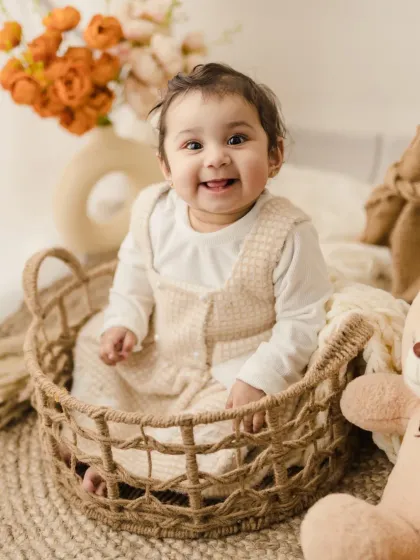 This little sweetheart is sitting in a basket with her teddy bear, giving us a warm and happy smile.