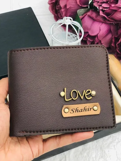 A close-up of a personalized brown leather wallet held in hand, featuring a 'Love' charm and an engraved nameplate.
