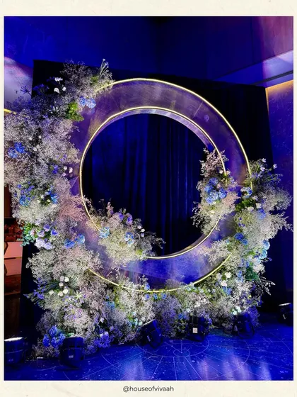 A modern and artistic photo backdrop featuring a glowing golden ring wreathed in an explosion of baby's breath and blue florals, set against a deep blue curtain.