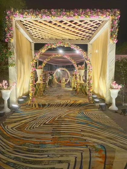 A beautiful evening entrance featuring a wooden trellis roof and floral arches leading into the venue. The combination of a uniquely patterned carpet and warm lighting creates a welcoming and stylish entryway.