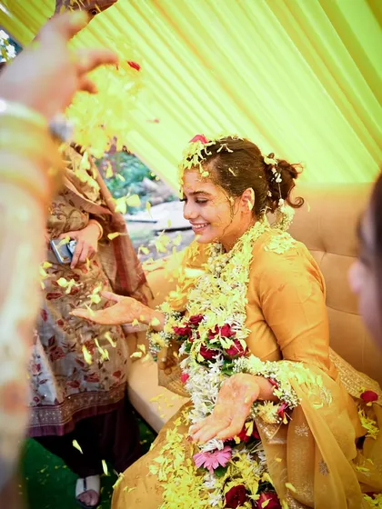 A candid shot of a bride enjoying her Haldi ceremony, showered with yellow flower petals. The image is full of movement, color, and genuine happiness.
