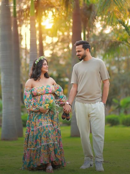 A romantic outdoor portrait of a couple holding hands and baby shoes. The golden light and floral gown create a warm and dreamy atmosphere.