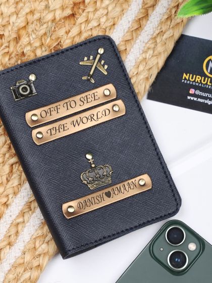 A black personalized passport cover with name, crown, camera, and airplane charms, plus "Off to see the world" quote tags.