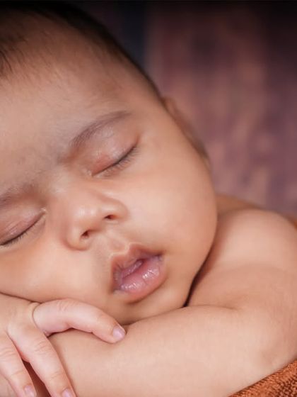 A close-up shot capturing the peaceful expression of a sleeping newborn. The soft brown wrap and simple background keep the focus entirely on the baby.