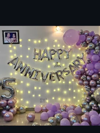 A beautiful 25th-anniversary decoration at home with a fairy light wall, 'Happy Anniversary' foil balloons, and a purple and silver balloon garland.