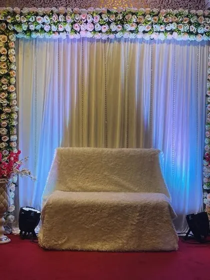 A classic backdrop for an engagement ceremony. The stage features a white curtain framed with artificial flowers and a comfortable sofa for the couple.