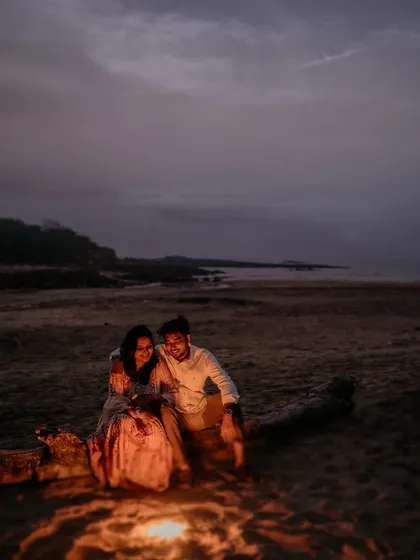 A cozy and unique pre-wedding shot of a couple by a bonfire on a beach at dusk. The warm glow of the fire creates an intimate and magical atmosphere.