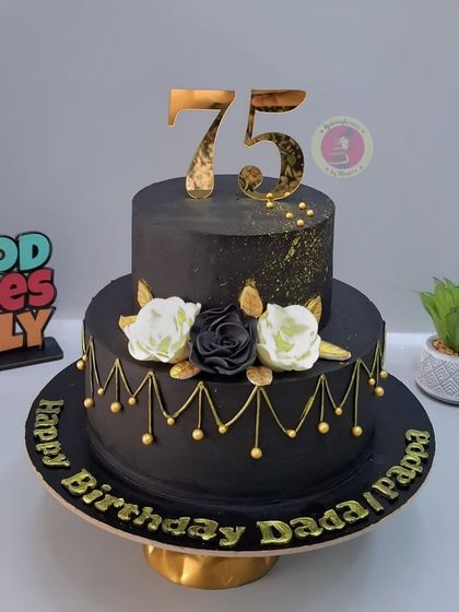 A grand two-tier black and gold cake for a 75th birthday, adorned with elegant black and white sugar flowers.