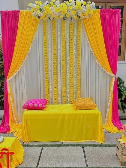 A clean and elegant Haldi stage setup with bright pink and yellow drapes. The backdrop is accented with vertical strings of white flowers, creating a simple yet beautiful space for the ceremony at home.
