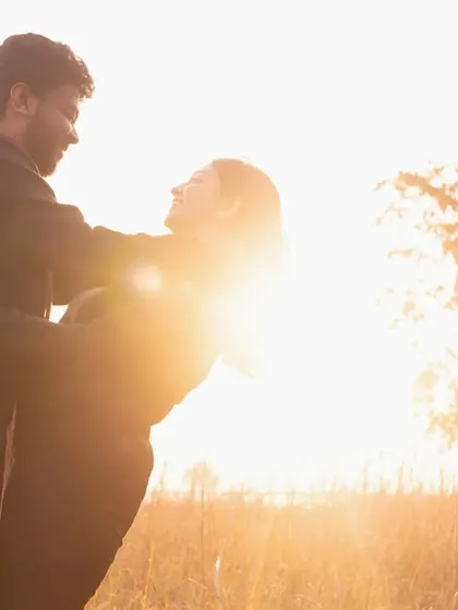 A playful and dynamic pose in the golden sunlight. He lifts her as they laugh together, creating a moment of pure, uninhibited joy against the backlit field.