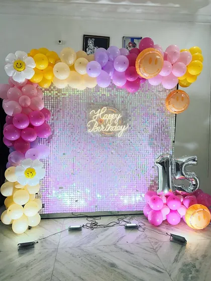 A fun and youthful birthday setup with a shimmering iridescent sequin wall. The decor includes a pastel balloon arch with smiley face and daisy accents, perfect for a 15th birthday party.