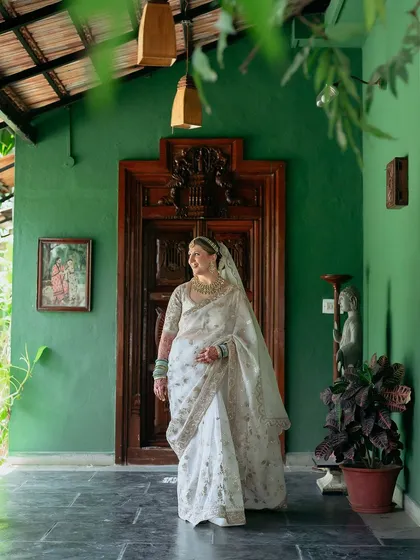 A bride in a beautiful white saree stands on the green-walled verandah, a picture of serene elegance.