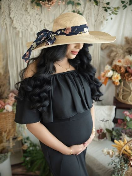 A chic and mysterious portrait of a mother-to-be in a black gown and a stylish sun hat. The hat adds a touch of glamour to the earthy boho setting.