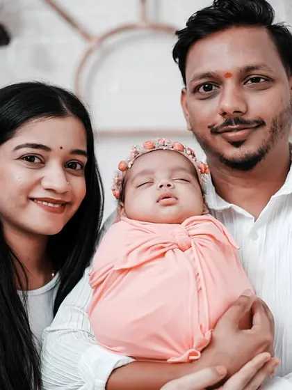 A close-up family photo, showing the parents' happy smiles as they hold their sleeping newborn daughter. Her floral headband adds a sweet touch.