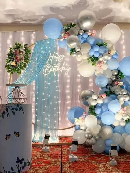 An elegant birthday setup with a blue-draped backdrop and fairy lights. The decoration includes a balloon arch in shades of blue, white, and silver.