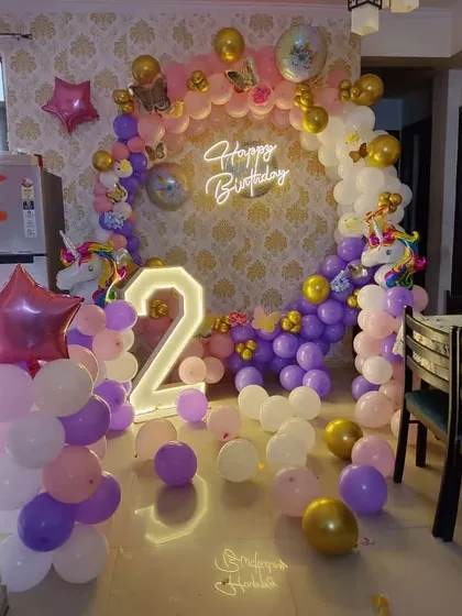 A unicorn-themed second birthday party inside a home. The setup includes a circular balloon arch with unicorn foil balloons, a neon sign, and a light-up number '2'.