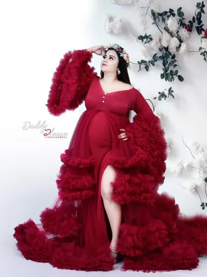 A high-fashion pose in a dramatic red ruffled gown. The mom-to-be looks up, showcasing the dress's incredible volume and the elegant floral backdrop.