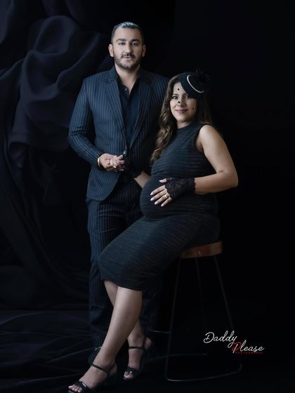 A classic couple's portrait in a dark and moody style. The parents-to-be are dressed in formal black attire, creating a timeless and romantic maternity photo.