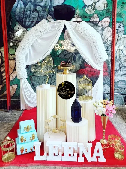 An elegant white-themed birthday setup with a fabric-draped arch. The arrangement of plinths and decorative items creates a sophisticated look.