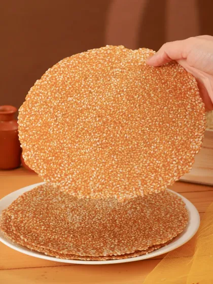 A shot capturing the thin, crispy texture of til-gajak, a traditional winter sweet. The hand element shows the product's delicate nature.
