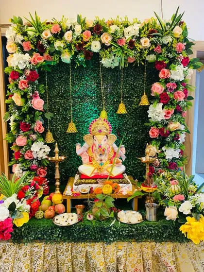 This home Ganpati setup combines a lush green backdrop with a frame of soft-colored roses and traditional bells, creating a fresh and vibrant look.