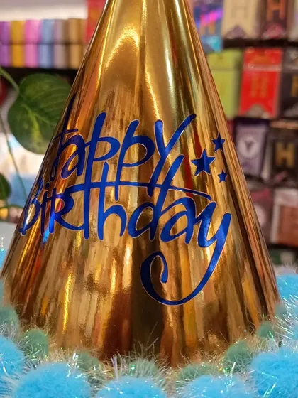 A close-up of the shiny gold birthday hat with blue text and pom-poms. A great accessory to make the guest of honor stand out.