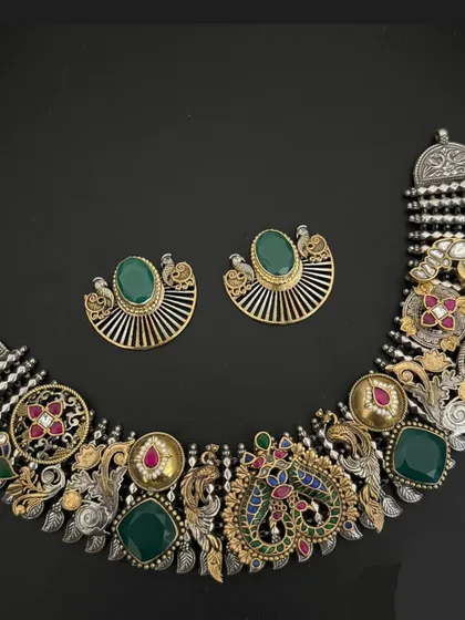 An intricate masterpiece from our fusion collection. This necklace set combines an oxidised silver base with gold-plated elements, green stones, and detailed peacock and floral motifs for a truly one-of-a-kind look.