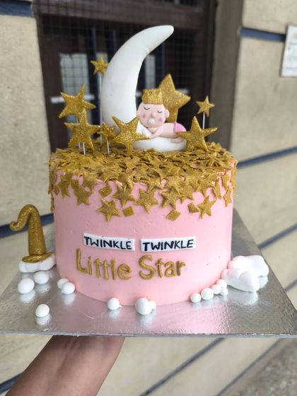 A "Twinkle Twinkle Little Star" themed cake for a baby's celebration. This pink cake is adorned with a sleeping baby on a crescent moon, surrounded by a cascade of golden stars.