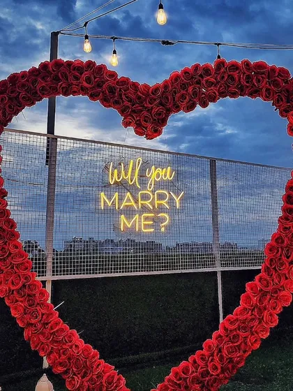 A close-up shot of the iconic red rose heart arch against a cloudy evening sky. The "Will you MARRY ME?" neon sign glows from within, highlighting the detail and craftsmanship of this classic proposal element.