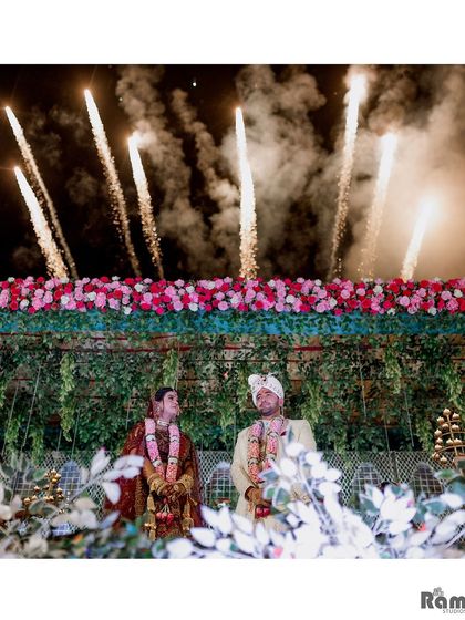 A stunning wide-angle shot of the couple on their wedding stage, with fireworks exploding in the sky above them, creating a truly spectacular image.