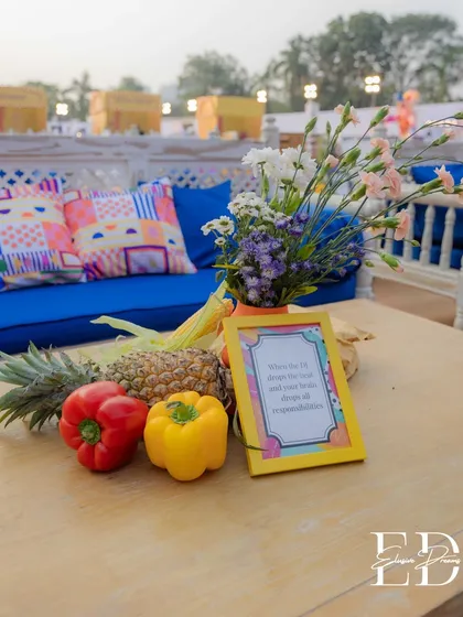 A sustainable and colorful centerpiece using fresh pineapples, bell peppers, and corn on a rustic wooden table. The bright blue sofa and patterned cushions in the background complete the lively carnival look.