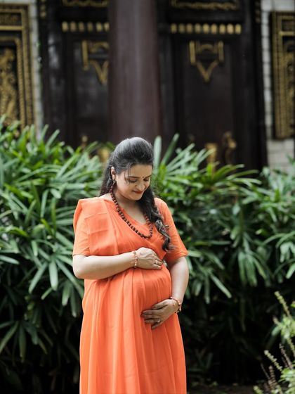 A serene solo portrait of the mother-to-be, dressed as Seetha. Her gentle hold on her baby bump against a backdrop of lush greenery and classic architecture creates a timeless image.