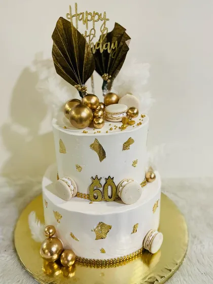 A grand two-tiered white and gold cake for a 60th birthday. This luxurious design is adorned with gold leaf, macarons, and dramatic fan toppers.