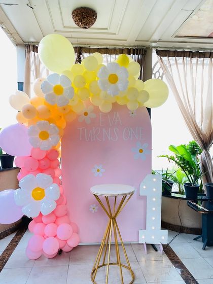 A cheerful daisy-themed first birthday party. The pink backdrop is adorned with a pastel balloon garland featuring cute daisy flower balloons.