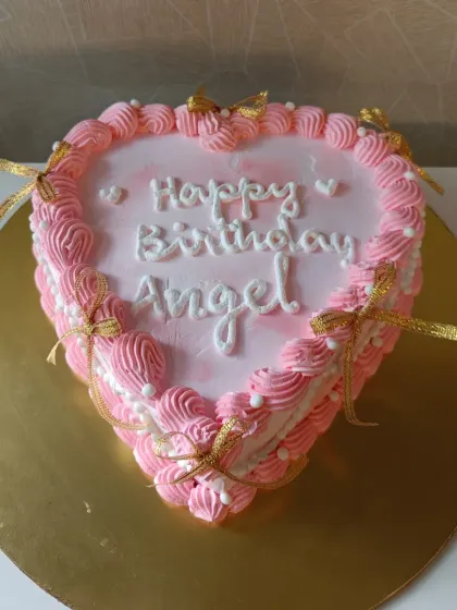 A sweet pink heart-shaped cake for an "Angel." This classic design features piped buttercream borders and delicate golden bows.