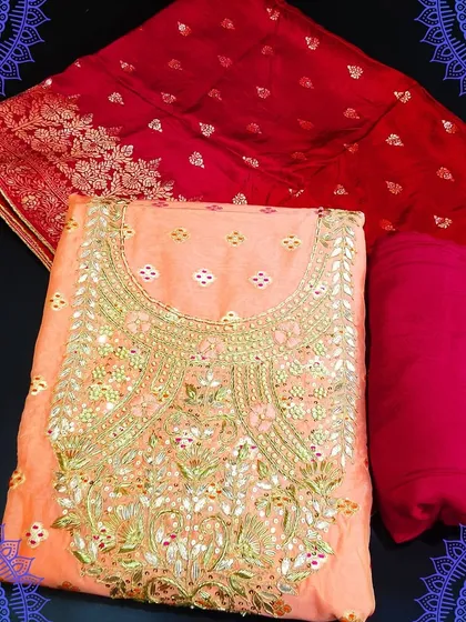 A lovely peach-colored dress material with intricate gold embroidery on the neckline. It comes with a rich red Banarasi dupatta, creating a beautiful contrast for a festive look.