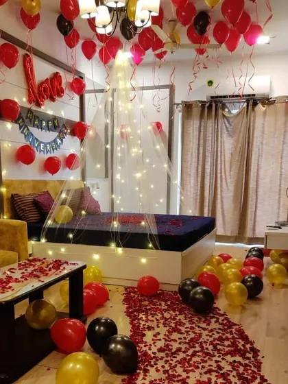 An elegant anniversary room decoration with a pathway of rose petals leading to the bed. The setup includes a fairy light canopy, balloons on the floor, and 'Love' foil balloons above the headboard.