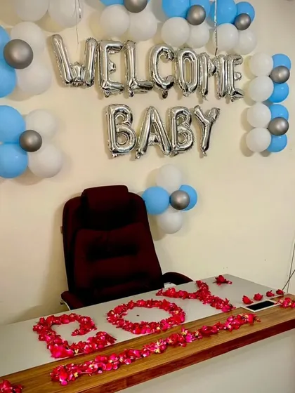 A classic "Welcome Baby" setup for a boy, with silver foil balloons and clusters of blue, white, and silver balloons. The table is decorated with rose petals spelling out "BOY" for a personal touch.